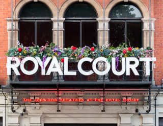 A woman walks beside the iconic front entrance of the Royal Court Theatre, its glass doors emblazoned with the words come in