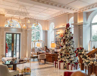 A frost-white Christmas tree with wrapped gifts adds festivity to the brightly-lit hotel lobby, seen over a banister garland.