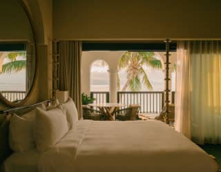 View over the white bed in an unlit room to the balcony with seating and sublime views of the sea, brushed by palm fronds.
