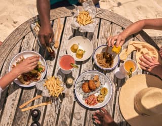 Diners' hands reach for grilled crayfish, corn, fries, and tasty plates at a Cap Shack table in the sand, seen from above.