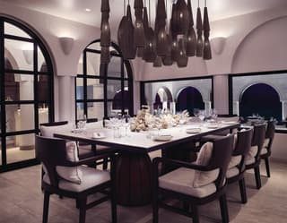 Ten dark wood chairs with white cushions are set around a white-topped dining table, beneath a cluster of grey pendant lights