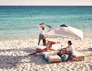 Four guests gathered on sunbeds under a white parasol on a sandy beach beside turquoise blue water