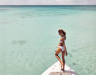 Lady in a white swimsuit balancing on the bow of a small boat in shallow clear waters