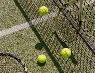 Detail of the tennis court, where a racket lies on the green turf and another rests on the net, with three discarded balls.