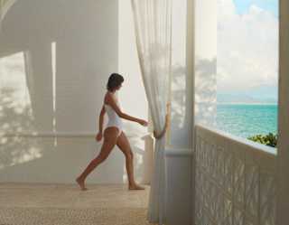 A woman in a white bathing suit walks across the cool spa floor to the terrace where the azure sea is glimpsed over a wall.