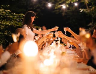Cut crystal wine glasses shine in candlelight as they are held aloft in a toast by a long table of diners in a night garden