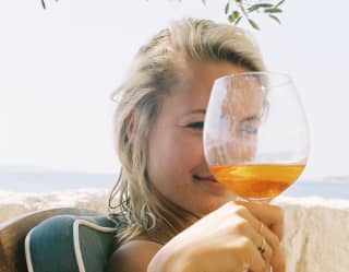 A young woman looks at the camera through the top of a glass of Aperol as she sits in the shade of an olive tree