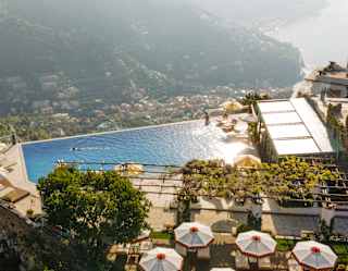 Aerial view of a deck with loungers and six parasols by a hillside infinity pool where guests enjoy breathtaking coast views.