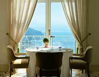 A round table set for two with beige chairs sits by a window with sheer curtains, offering a view of the sea and coastline under a clear sky. A small potted plant decorates the table.