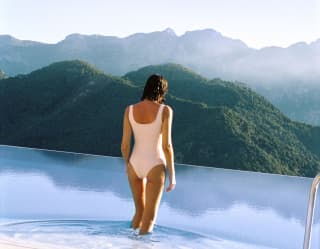 A woman in a white swimming costume steps into the cool blue infinity pool with a verdant hilly backdrop, seen from behind.