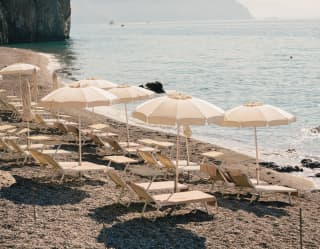 Two pristine rows of chaise longues lie facing the sea on a private pebble beach, each pair shaded by a white parasol