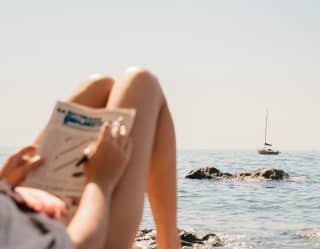 Lady on a beach sunbed reading a paper with a fishing boat bobbing on the waves beyond