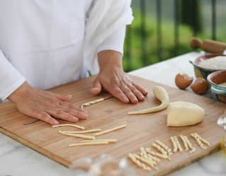 Close-up of Mamma Felicia's hands as she demonstrates how to make busiate pasta on a wooden board with dough and a ferretto.