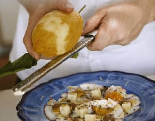 A chef grates the zest of a large lemon onto a salad plate of cubed cheeses, mandarin segments and olives