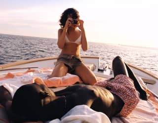 A woman raised up on her knees photographs her lounging companion as they relax on towels on the deck of a private boat.