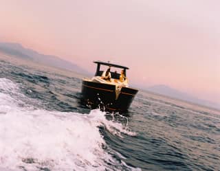 Angled view over the choppy wake of a boat as it carries two guests, seated on the back deck, towards a pink hazy sunset sky.