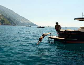 A woman dives from the back deck of a boat as her male friend canon-balls, both captured in mid-air, with an ocean backdrop.