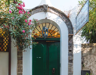 Green door in an arched doorway surrounded by potted plants and pink hanging flowers