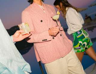 Close-up of two men from the shoulders down enjoying poolside cocktails, as a woman in a green-patterned skirt passes behind.