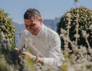 Chef Armando Aristarco wears long sleeve chef whites and smiles as he picks fresh oregano from the hotel gardens