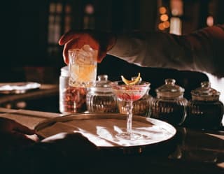 A hand is about to place a signature cocktail, Onça Pintada on a serving tray next to a pretty pink drink in a crystal glass.