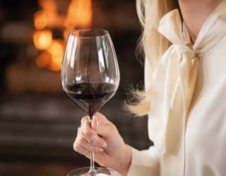 A smiling blonde woman in white trousers and blouse holding a large glass of red wine in front of an open fire