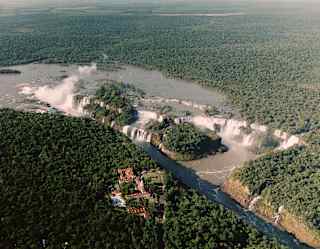 Aerial view of Hotel das Cataratas in forest opposite the vast ledges and pools of the Iguassu falls with plumes of spray.