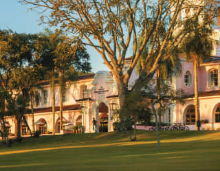 Sun shines between exotic trees onto the period terraced façade and lawns of the Hotel das Cataratas