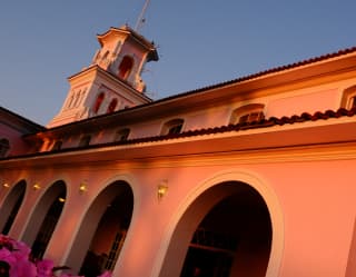 Angled image of an arcade walkway and main hotel behind, with its tower rising into blue sky, seen over pink geraniums.