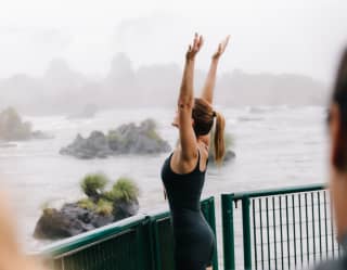 At a viewpoint, a woman in black Lycra and a pony tail faces the Iguassu Falls with arms stretched high, feeling the spray.