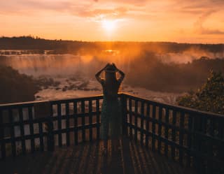 Lady in a green dress and sunhat standing on a platform overlooking the Iguassu Falls in an orange sunrise.
