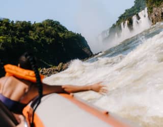 A woman in a lifejacket reaches from a Macuco Safari boat to feel the river spray, seen from behind with the falls up ahead.