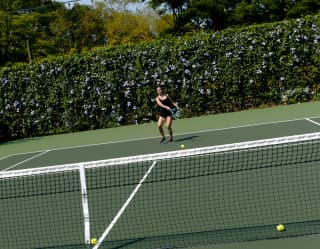 Angled image of nine yellow balls on a dark green tennis court, where a woman in black practices shots from across the net.