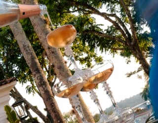 Angled image of a three-tier pyramid of coupe glasses, with pink champagne pouring into the top one, at an al fresco event.