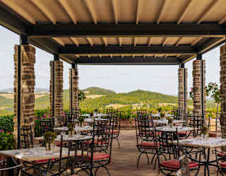 Stone columns support a canopy above two rows of four tables awaiting guests at Emporio Terrazza, with Tuscan hill views.