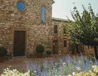 A rustic stone building with arched windows and a wooden door stands under a bright, clear sky. Lavender and green shrubs line the foreground, and a leafy tree is visible on the right.