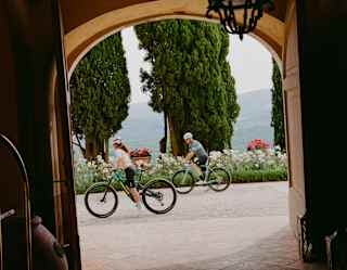 Two people ride bicycles past an arched doorway framed by tall trees and flowers, with mountains visible in the background. The scene is viewed from inside a shaded entrance.