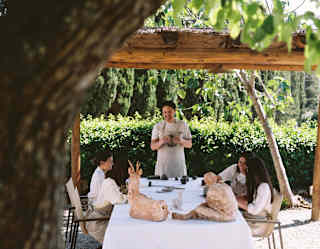 In an open outdoor structure, four guests in beige aprons attend a clay workshop at a table with sculpted figures.