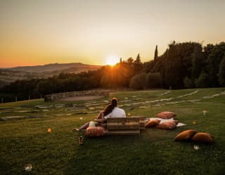 Seen from behind, a cosy couple watches the sun set from the amphitheatre lawn, surrounded by cushions and picnic baskets.