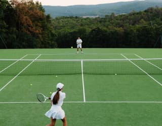 High-angle view of a ball-strewn tennis court, as a woman in the foreground hits a ball over the net towards a male opponent.