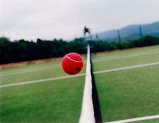 A red tennis ball, captured in a still as it crosses the net in an eye-level view of the lush green tennis court.