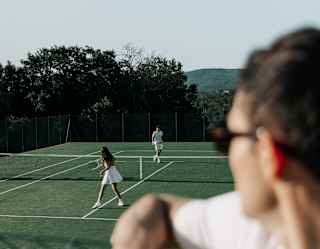 A person watches a tennis match from behind a fence, with two players in white on a green outdoor court under a clear sky. Trees and hills are visible in the background.