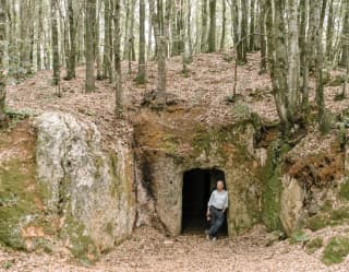 A man leans casually against the entrance to an ancient Etruscan tomb, almost hidden in a woodland hollow