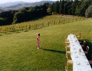 On the lawn, a woman in a red and white towel gazes at the view as she stands near a long celebration table, seen from above.