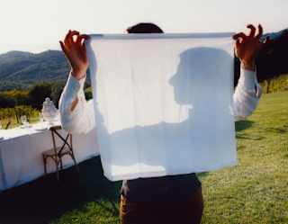 A man holds up a white napkin which shows the shadow of his face in profile, seen at a celebration table in the garden.