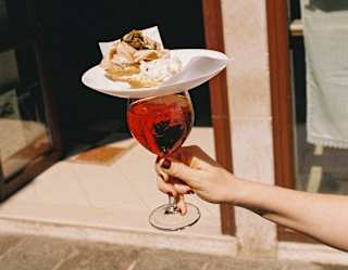 A plate of a cicchetti pastry treats rests on the top of a glass of iced Aperol, carried in a Venice street, seen close-up.