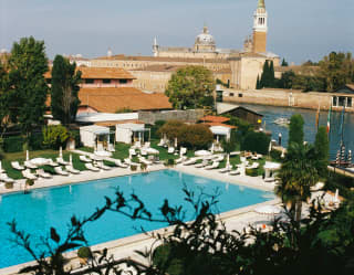 Chairs and folded-down parasols line the sapphire hotel pool, overlooked by stone and terracotta buildings, seen from above.