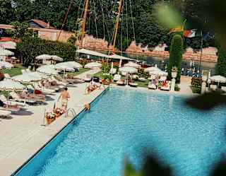 Boat masts rise from the canal behind the pool in a view capturing guests relaxing below parasols, part-obscured by foliage.