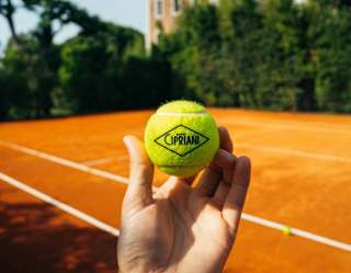 A man's hand holds a Cipriani-branded yellow tennis ball, with the clay-surfaced court and hotel in the background.