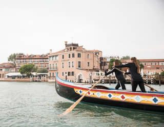 Two women practise the art of Venetian Rowing, standing and facing forward in a large gondola-style colourful boat